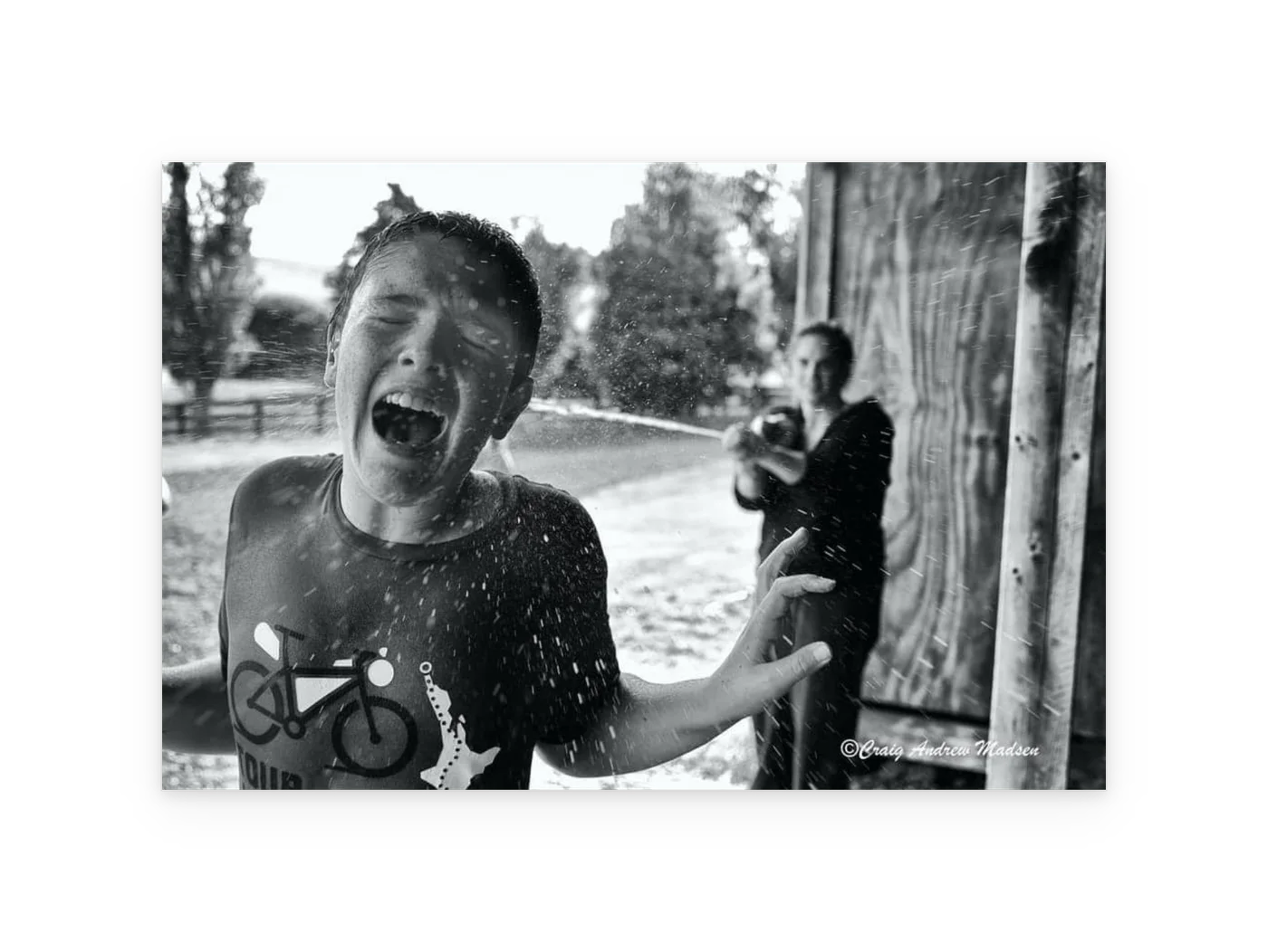 An unframed black and white print of a boy laughing as he is sprayed with a hose.