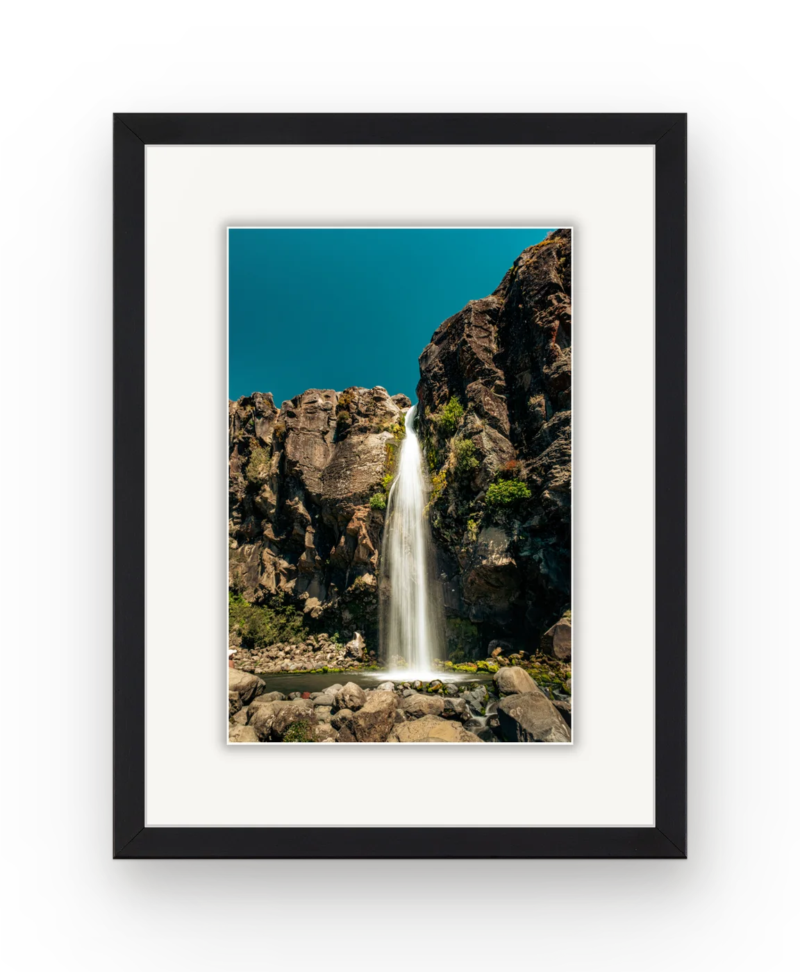 A waterfall falling down a dark rock face, framed in black with a white mat.
