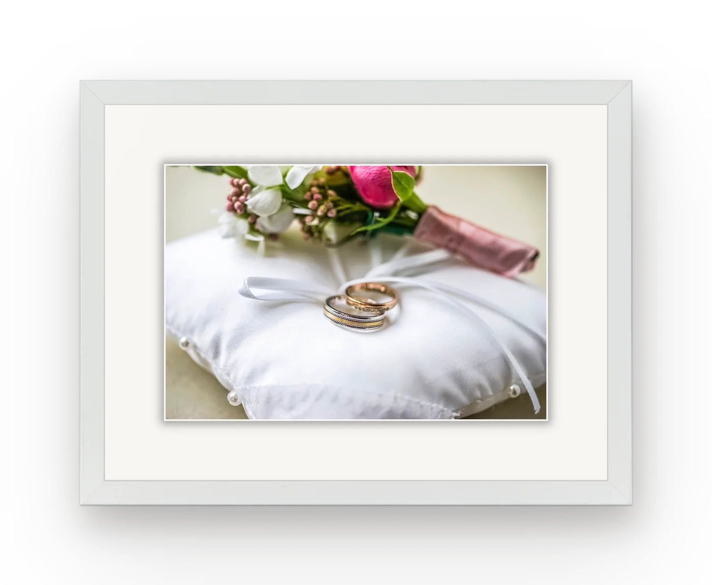 Two wedding rings on a satin ring pillow, framed in white with a white mat.